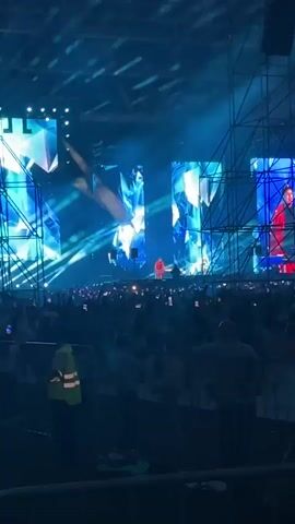 A concert is taking place in a large indoor venue. The stage is illuminated with bright blue lights and features large LED screens displaying abstract, geometric patterns. Several performers are on stage, dressed in colorful costumes. A large crowd of people is visible in the foreground, many holding up their phones to record the event. A security guard in a high-visibility vest is standing near the front of the crowd. The overall atmosphere appears to be energetic and celebratory.