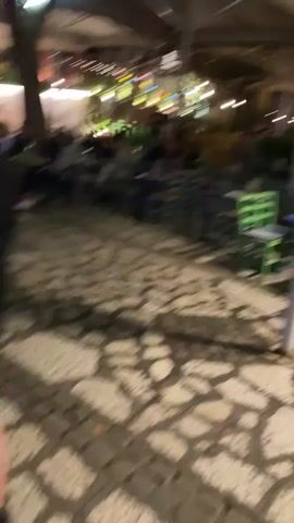 The video depicts an outdoor café area at night, with patrons seated at tables and chairs of various colors. The ground is paved with stone. A large umbrella provides cover over part of the seating area. Beyond the café, a street with a sidewalk, small buildings, and a "STOP" sign can be seen. A few people are walking through the café area.