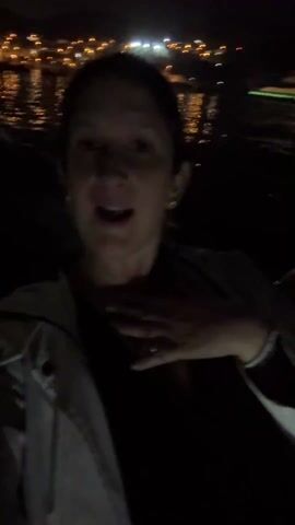 A woman in a black top and a white jacket is filming a selfie video at night. She smiles and poses for the camera, occasionally touching her chest. In the background, there's a body of water with city lights reflecting on its surface.