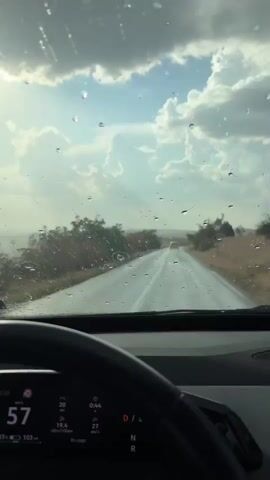 The video is filmed from inside a car, looking out through the windshield. Raindrops are visible on the windshield, and the wipers are activated briefly. The car is driving on a wet asphalt road through a rural landscape with sparse vegetation and trees. The sky is cloudy, with patches of sunlight breaking through. The dashboard of the car is partially visible, showing a digital display with speed and other information, with the current speed indicated as 57 km/h. In the distance, another car is visible on the road.