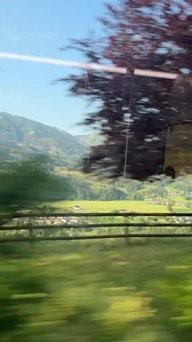 The video shows a view from a moving vehicle, likely a train, traveling through a lush green valley with mountains in the background. The foreground is blurred due to motion, with trees and a wooden fence visible. The landscape transitions from a close-up of trees to a wider view of a village nestled in the valley, surrounded by green fields and forests. The sky is clear blue with a few wispy clouds. The overall impression is of a scenic journey through a rural mountainous region.