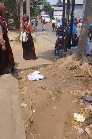 The video shows a group of people, many of whom appear to be students in uniform, gathered along a street on a sunny day. In the foreground, two individuals in brown uniforms with headscarves stand on a concrete path. One is facing left, with a backpack over their shoulder. The other is facing away, holding a phone. Scattered on the ground in front of them are pieces of paper and trash.

Further down the street, a larger group of people are visible. Many are wearing blue and white uniforms, some with hats. One person is sitting on a low concrete structure on the right side of the frame, wearing a yellow jacket and a helmet, with a water bottle next to them. A red and black motorcycle is parked near a utility pole. In the background, a white van is parked on the street, and several houses are visible. The overall impression is an outdoor gathering or event, possibly a school activity or a parade, taking place in a somewhat unkempt urban or suburban environment.