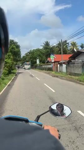 A first-person perspective video shows a person riding a blue scooter on an asphalt road with a white dividing line. On the right side of the road are houses with fences and greenery. A white bus with blue and yellow stripes is approaching. The sky is blue with white clouds. The scooter's side mirror shows a reflection of the rider wearing a helmet. Overhead electrical wires are visible.