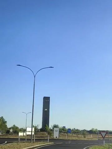 A wide shot captures a clear blue sky above a flat landscape. In the foreground, a two-headed street lamp stands tall on the left side of the frame. To the right of the street lamp, a tall, dark, rectangular structure, possibly a monument or tower, is visible in the distance. In front of the tower, a small white sign with text and a logo is present. A road curves through the scene, with white markers along its edges. In the middle ground, there are some trees and a patch of cultivated land. Further in the distance, the landscape appears to be agricultural fields. A few other signs are scattered throughout the scene, including a blue directional sign with an arrow and a triangular road sign on the far right. The overall scene suggests an outdoor, possibly rural or semi-rural, environment under a bright, clear sky.