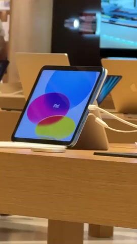 The video shows the interior of a brightly lit electronics store, likely an Apple Store, with a minimalist design featuring light wood tables and a white tiled floor. The camera is initially focused on a tablet display on a wooden table. The tablet is a light blue iPad, displaying a colorful wallpaper with circles. Several other iPads and laptops are visible on the table and other tables in the background. The store appears to have multiple rows of display tables, with customers browsing. In the background, there are shelves with more electronic devices and large screens displaying content. The overall atmosphere is clean, modern, and spacious. The camera pans slowly to the left, revealing more of the store's layout and the people within it. Several individuals are seen interacting with the displays or walking through the store. The ceiling has a distinctive slatted design with embedded lighting. The video captures a typical retail environment where customers can explore and try out electronic products.