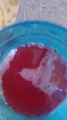The video shows a close-up of a blue plastic cup filled with a red, bubbly liquid, likely a beverage. Next to the cup is a piece of white food, possibly a bun or bread, on a brown paper. The background is a blurry gray surface. The camera angle is from above.