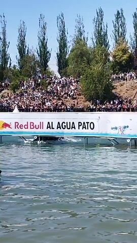 The video shows a water sport event taking place in a lake or river. The event appears to be sponsored by Red Bull. Spectators are watching from a hillside. A person on a jet ski is riding on the water, with a paddleboard attached. Another person is swimming in the water.