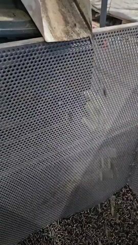 The video shows small black pellets falling from a metal chute onto a perforated metal surface. The chute is stained and appears to be part of some kind of industrial machinery. The pellets are cylindrical and appear to be uniform in size. The perforated surface is also metal and has a grid pattern. In the background, there are large white bags and some wooden pallets, suggesting an industrial or warehouse environment. The pellets are falling continuously throughout the duration of the video.