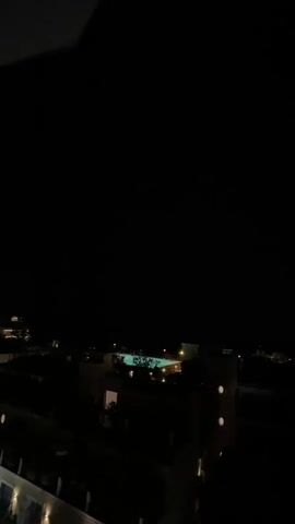 The video captures a night scene from a high vantage point, looking out over buildings and a swimming pool. The sky is dark initially, then a bright flash of lightning illuminates the scene with a purple hue, revealing the outline of clouds and a jagged bolt of lightning stretching across the sky.
