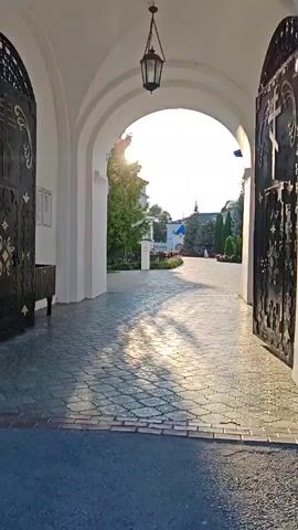 The video shows a view through an arched entryway, looking out onto a paved courtyard. The sun is shining brightly, casting shadows on the cobblestone ground. On the left side of the entryway, there is a black metal gate with decorative elements, and on the right, another similar gate is open. Inside the entryway, a dark colored trash can is visible. The courtyard outside has greenery, trees, and some buildings with blue roofs. The overall scene appears to be at a religious site, possibly a monastery or church, given the architecture and the gates. The light suggests it is either early morning or late afternoon.
