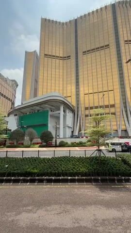 The video shows a street in Macau, China. The camera pans to the right to show the Galaxy Macau resort, a large complex of hotels and casinos. The buildings are tall and have a gold exterior. There are trees and other landscaping along the street. A few cars pass by on the road. The sky is blue with some white clouds.