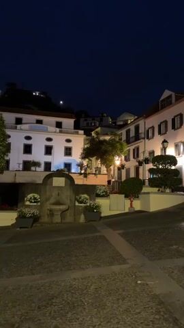 The video depicts a quiet, cobblestone town square at night. The sky is a deep, dark blue. The buildings surrounding the square are white with dark shutters and terracotta roofs. They are illuminated by warm streetlights. One building has a small balcony on the second floor. In the center of the square, there is a small fountain with white flowers planted around it. A neatly trimmed tree and bushes are planted in a raised section of the square. A motorcycle is parked near one of the buildings. There is a fire hydrant near the tree. A person walks in the bottom right corner of the frame. The sound of a person speaking is audible in the background.