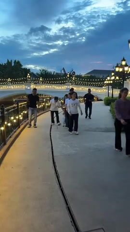 A group of people walk along a paved path next to a railing decorated with lights. The sky is dark blue with clouds. A bridge with lights and buildings are in the background.