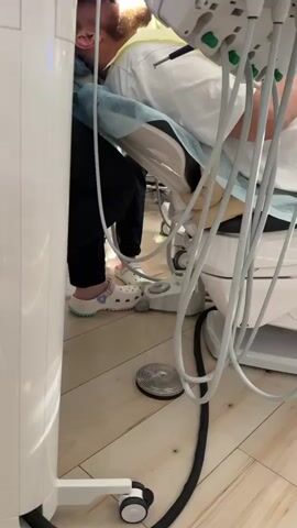 The video shows a dental procedure in progress. Two dental professionals, wearing masks and gloves, are working on a patient who is reclined in a dental chair. One professional appears to be the dentist, positioned closer to the patient's head, while the other, likely a dental assistant, stands beside them. They are using dental instruments, and a microscope is positioned above the patient, suggesting a detailed or complex procedure. The environment is a dental operatory, indicated by the dental chair, equipment such as a dental unit with various tools, and the general clinical setting. The lighting is bright and focused on the treatment area. The floor appears to be wooden.