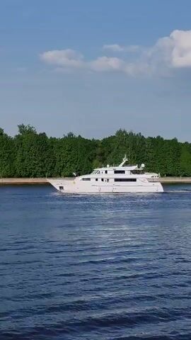 A white motor yacht is seen moving across a body of water, likely a river or wide canal, on a bright, sunny day. The water is a deep blue, with slight ripples indicating movement. The yacht is modern in design, with multiple decks and a clean, streamlined appearance. Lush green trees line the bank of the waterway, providing a stark contrast to the white yacht and the blue water. The sky is light blue with scattered white clouds.