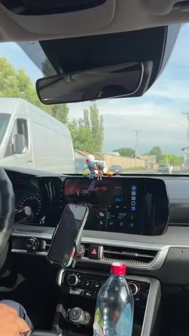 A first-person perspective video shows the interior of a car driving on a paved road. The dashboard features a large infotainment screen displaying a speed of 38 km/h and various icons. Two small figurines of a couple kissing are placed on top of the dashboard. A smartphone is mounted on a car phone holder, also on the dashboard. A bottle of water is visible in the center console. Outside the car, trees and buildings line the road, and other vehicles are present. The sky is cloudy. The video begins with the car moving at 38 km/h and the speed increases to 42 km/h by the end.