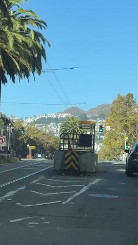 A gray SUV drives down a city street, passing a palm tree and a mountain in the background. The sky is clear and blue. There are some power lines visible overhead. The street has a crosswalk and traffic lights.