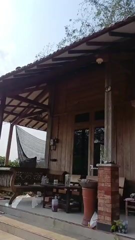 The video shows the exterior of a wooden house. The house has a covered porch with wooden beams and a tiled roof. There are several bird cages hanging from the porch. The porch is furnished with wooden chairs and tables, as well as some potted plants and water bottles. A security camera is visible on the porch. The background consists of trees and foliage.