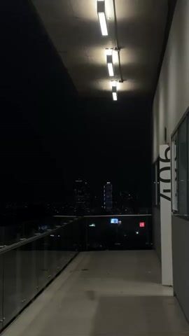 The video shows a balcony at night. A glass railing is visible on the left, and a concrete floor. Above, a series of lights are mounted on the ceiling. In the distance, city lights and buildings are visible against the dark sky. A sign with the number 904 is mounted on a wall to the right. The camera is stationary and the scene is dark.