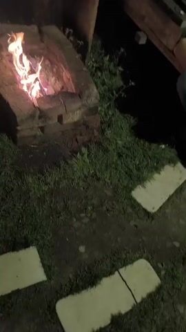 A video shows a fire pit with flames burning in the center. The fire pit is made of bricks and is surrounded by grass. In the background, there is a wooden fence and a pile of firewood. Several people are visible, sitting or lying on the grass. One person is wearing a green shirt, and another has on a gray and white patterned shirt. A person on the left is wearing red sneakers.