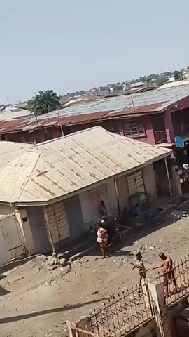 The video depicts an outdoor scene, likely a residential or commercial area, with visible signs of wear and tear. Several people are present. A shirtless man wearing dark jeans stands near a fence and appears to be interacting with a young girl wearing a colorful patterned shirt and a hair bun. Closer to the buildings, there are other people, including a woman in a patterned dress standing near a parked motorcycle. The buildings in the background appear to be old and have rusted corrugated iron roofs. The ground is uneven and covered with rubble and debris. The video is filmed from an elevated perspective.