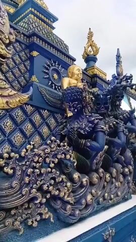 A close-up shot pans across a detailed blue and gold sculpture of figures. The figures are humanoid with demonic features, including horns and skeletal faces. They are adorned with necklaces made of skulls and appear to be in a praying or devotional pose. In the background, a golden statue of Buddha is visible, partially obscured by the demonic figures. The sculpture is elaborate, with intricate carvings and metallic accents. The environment appears to be outdoors, with a slightly overcast sky.
