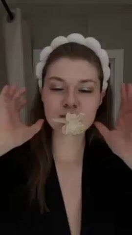 A woman in a black robe and a white puffy headband stands in a bathroom. She holds a small white flower in her mouth and then removes it. She holds the flower in her hand and looks at the camera. A shower curtain and white door are in the background. There is music playing.