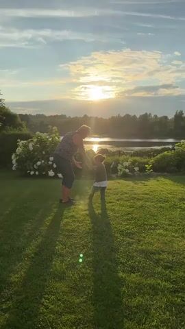 A person is interacting with a small child on a grassy lawn during sunset. The sun is low in the sky, casting long shadows. There is a body of water and trees in the background. The person is wearing a striped shirt and dark pants, and the child is wearing a green shirt and white pants. They are holding hands and appear to be walking together. The scene is peaceful and idyllic.
