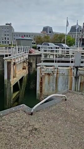 The video shows a lock gate mechanism. Water can be seen flowing through the gap between the two gates. A concrete walkway is in the foreground with metal edging. In the background, buildings and parked vehicles are visible. The sky is overcast. There are no people visible in the video. The gates appear to be made of metal and wood, with some signs of rust and wear. The water level appears to be low. The overall scene suggests a functional lock system in a maritime environment.
