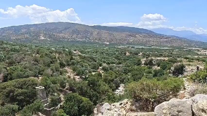 The video shows a drone flying over a hilly, arid landscape with sparse vegetation consisting of trees and bushes. The ground is rocky and uneven. In the foreground, there are some ruins, possibly stone structures, partially covered by vegetation. In the middle ground and background, the landscape is dominated by rolling hills covered in greenery, with more distant, larger mountains under a clear blue sky with some scattered clouds. The overall impression is of a rural, undeveloped area with signs of ancient human presence.