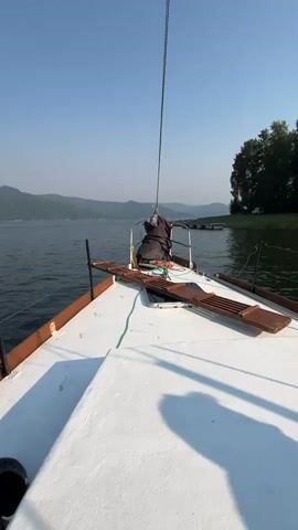 The video shows the view from the bow of a sailboat moving across a calm body of water. The water is dark and reflects the clear blue sky. On either side of the water are gently sloping hills covered in trees. The hills in the distance are hazy. The sailboat has a white deck with wooden trim. There is a person sitting on the deck near the front of the boat, wearing dark clothing. They appear to be leaning over a railing. A rope runs up from the bow to the top of a mast, which is not fully visible. In the distance, there are docks or moorings extending into the water. The sun is shining, casting shadows on the deck.