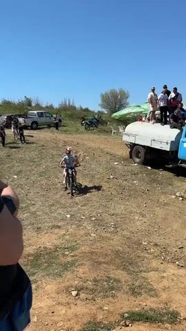 The video depicts two boys on a black motorcycle in an outdoor setting. The boy in front wears a black cap, light blue t-shirt, and black shorts. The boy behind wears a red and white cap, gray t-shirt, and blue shorts. They are sitting on the motorcycle, which is parked on a dirt ground. Behind them is a blue truck with people standing on its tank. Other vehicles and people are scattered in the background, under a clear blue sky. The overall atmosphere is relaxed and casual.