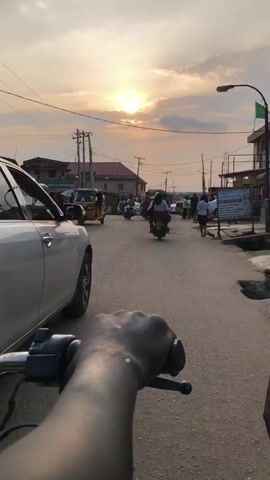 The video begins with a close-up shot of a person's hand gripping a motorcycle handlebar. The perspective is that of someone riding the motorcycle. The camera then pans to show a street scene bathed in the warm light of a sunset. Several motorcycles and a few cars are visible on the asphalt road. Pedestrians are walking along the side of the road, and some are interacting with vendors. Buildings with various signs and awnings line the street, suggesting a commercial or residential area. Power lines crisscross the sky. The overall atmosphere appears to be that of a busy street at dusk.
