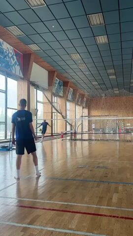 This video shows a person playing badminton in an indoor gymnasium. The person, wearing a blue t-shirt and black shorts, is positioned on the right side of the court, facing the net. They are in the process of hitting a shuttlecock. The gymnasium features a wooden floor with court markings and a high ceiling with a grid of square panels. A net stretches across the middle of the court, separating two sides. In the background, there are large windows and more sports equipment, including what appear to be basketball hoops. The lighting suggests it is daytime.