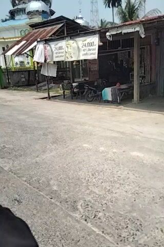 The video shows a street view with a building and a car. In the foreground, the street appears to be paved with concrete. To the left, there is a building with a tin roof and a colorful facade. Signs with Indonesian text and prices are visible. A motorcycle is parked near the building. Further down the street, a mosque with green and white accents is visible. A tree stands in front of the mosque. A black SUV is parked on the right side of the street, partially obscuring the mosque. The ground is uneven in some areas, with cracks and debris. The overall impression is of a street scene in a residential or commercial area.