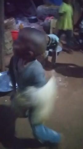 The video shows three young children in what appears to be an outdoor setting, possibly a yard. The primary focus is on the child in the foreground, who is wearing a dark-colored shirt and blue pants, and appears to be holding onto a light-colored, possibly furry or feathery object or garment. The child is looking towards the camera, smiling and making facial expressions. In the background, another child is wearing a light blue t-shirt with a panda graphic on it and looking directly at the camera. A third child is seen to the right of the first child wearing a green shirt. The background is cluttered with various objects, including a red bucket, logs of wood, cooking utensils, and what appears to be basic furniture. The overall lighting is dim, and the video appears to be taken at night.