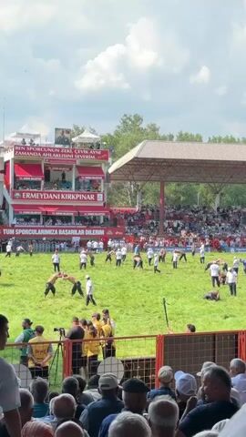 The video shows an outdoor wrestling event. In the foreground, a crowd of spectators watches from behind a fence. In the mid-ground, wrestlers are engaged in matches on a grassy field. There are several pairs of wrestlers actively grappling. In the background, a large grandstand filled with spectators is visible, with banners and a large screen. The event appears to be a formal competition, possibly a tournament, given the organized seating and the number of participants. The environment is sunny with some clouds in the sky.