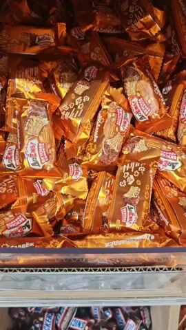 The video shows a close-up view of different types of candy. The first section displays an abundance of candies with shiny orange wrappers, featuring designs of nuts and what appear to be grains. These candies are piled high in a container. The second section shifts to a view of numerous Snickers candy bars, all individually wrapped in their characteristic brown packaging with blue and red lettering. Amongst the Snickers, a single candy with a purple and green wrapper is visible. The third section shows a pile of round candies wrapped in golden foil. The final section reveals a large quantity of candies with vibrant purple and lime green wrappers, adorned with star-like patterns and Cyrillic text.