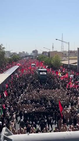 This video shows a large procession of people carrying many red flags and some green and white flags. The crowd is very dense and extends far down a wide street. In the center of the procession, there are at least two trucks, one in front of the other. People are walking, and some are standing on top of the trucks. The environment appears to be an urban area with buildings visible in the background, and the sky is clear and blue, suggesting it is daytime. The overall impression is of a significant gathering or demonstration.