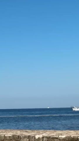 A wide shot captures a tranquil ocean under a clear blue sky. The water is a deep blue, with a subtle ripple on its surface. A white boat with yellow accents is visible in the distance, moving horizontally across the frame from right to left. It appears to be traveling at a moderate speed. In the foreground, a stone wall with a rough texture runs horizontally across the bottom of the frame, providing a sense of foreground context. The overall scene is peaceful and serene, suggesting a calm day at sea.