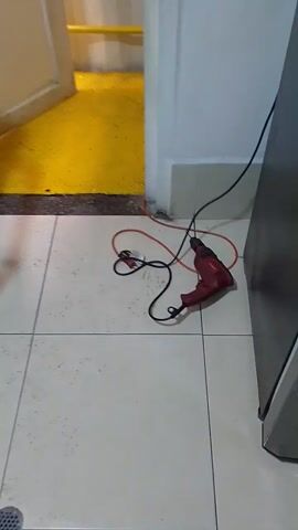 The video shows a red power drill lying on a tiled floor. The drill has a black power cord and an orange extension cord plugged into it. The floor is white with light grey grout.  A yellow grated surface and a white cabinet are visible to the left of the drill. Above the drill is a gray metallic surface, possibly a cabinet.  The video is shot from above.