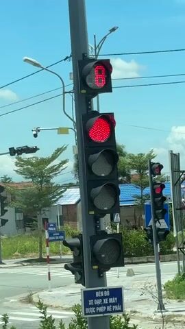 A traffic light pole stands tall against a blue sky with scattered clouds.  The traffic light itself has a digital display showing a countdown timer, starting at 7 and decreasing to 3. Above the timer, a red light is illuminated, and below it, a larger red circle is also lit.  Further down the pole are several unlit black circular traffic light lenses. In the background, there are trees, buildings with blue roofs, and other traffic signals. A white sign with blue text is visible at the base of the pole.