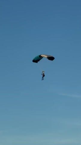 A lone skydiver with a blue and grey parachute descends against a clear blue sky. The skydiver is wearing a dark jumpsuit and helmet. The parachute is open and appears to be functioning normally, guiding the skydiver's descent. The camera is looking up at the skydiver from below.