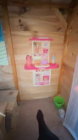 The video opens showing a toy kitchen set mounted on a wooden wall. The camera pans to the left, revealing a wooden shelf with a baby doll and various toy dishes, including a red cup, a yellow bowl, and blue plates. A piece of paper with writing on it is also visible on the shelf. The camera then moves to the right, showing a green bucket and other items on the floor. Finally, the video focuses on the piece of paper on the shelf, which appears to be a handwritten menu. The overall environment is a dimly lit room with wooden walls and a carpeted floor.