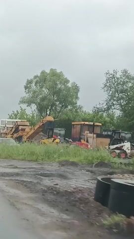 The video appears to be a compilation of several short clips taken from a moving vehicle. The overall scene depicts an outdoor industrial or construction area, likely in a rural or semi-rural setting, possibly Kabanikha, Russia.

The first clip shows heavy machinery, including a large yellow conveyor belt system and what looks like a truck or vehicle. The area is overgrown with tall green grass and foliage. There are also some wooden structures or containers in the background. The sky is overcast, suggesting cloudy or rainy weather.

The second clip is very blurry and shows a similar scene with heavy machinery and greenery, but the motion of the vehicle makes details difficult to discern. The weather appears to be poor, with significant blurring likely due to rain or spray.

The third clip shows a vast landscape with large piles of sand or gravel stretching into the distance. A road or path is visible in the foreground, with tire tracks indicating vehicle use. The sky is heavily overcast, and the overall impression is one of a working industrial site under a grey sky.

There is no discernible text in any of the clips. The time of day appears to be daytime, given the natural light, but the overcast weather obscures any specific indications of morning, afternoon, or evening. The overall mood is functional and industrial, with no human presence directly visible. The activity taking place, based on the machinery and materials, is likely related to construction, material processing, or excavation.