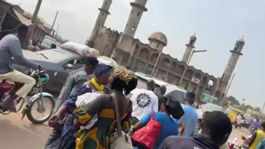 The video shows a busy street scene in front of a large mosque. The mosque has three minarets and a multi-tiered facade. Several people are walking and standing around, engaged in conversation and movement. There are cars and motorcycles on the street, indicating a busy traffic environment. Some of the cars have goods piled on their roofs. The scene takes place during daylight under a moderately overcast sky.  The general impression is that of a market day near a significant landmark.