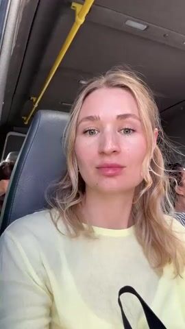 A blonde woman with green eyes and light skin is sitting in a bus seat, looking directly at the camera. She is wearing a pale yellow crew neck sweater. Her hair is wavy and falls to her shoulders. She is wearing cross-shaped earrings. Behind her, another passenger is visible in a striped shirt. A yellow grab bar runs along the ceiling of the bus. The lighting suggests it is daytime.