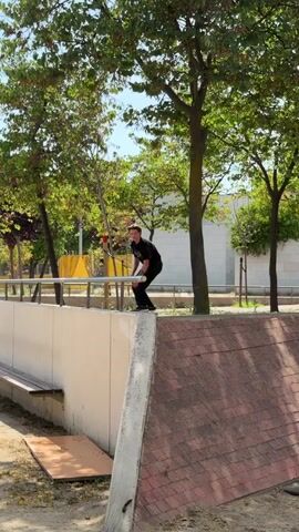 A person on a skateboard is performing a trick in a park. They skate towards a ledge, grind on the ledge with their skateboard, and then jump off the ledge. They land on the ground and continue to skate. The park has trees, benches, and a concrete structure. The person is wearing a black t-shirt and black pants. They are in mid-air with their arms outstretched. The skateboard is metallic and has wheels. The ground is covered with dirt and some leaves. The sky is visible with some clouds.