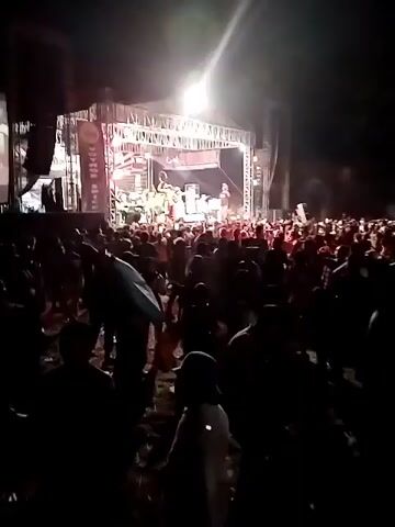The video shows a nighttime outdoor concert. A band is performing on a stage under bright lights. The band appears to be a group of musicians playing various instruments, possibly including keyboards and percussion. Several singers or performers are on stage, with some dressed in traditional or colorful clothing. A large banner above the stage displays the name "Com Lorensa". In front of the stage, a dense crowd of people are gathered, many of whom are holding up their phones to record or take pictures. The crowd is dark, with some individuals visible in silhouette. The overall atmosphere suggests a lively and energetic event.