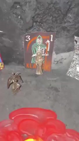 The video shows an altar with a red candle surrounded by melted wax. In the background is a picture of the Grim Reaper next to a silver-colored miniature of a robed figure and a figurine resembling a gargoyle or winged demon. The altar sits against a dark, textured backdrop, possibly meant to represent a cave or rocky environment. The camera then moves to show a tattoo on someone's skin, which appears to depict the Grim Reaper figure from the altar in the background.